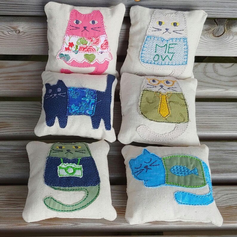 May include: Six handmade cat-shaped pillows with different designs. The pillows are made of white fabric with embroidered cat faces and details. The cats are wearing different outfits, including a pink dress, a blue shirt, a green tie, a blue shirt with a camera, a blue shirt with a fish, and a white shirt with the word "MEOW".