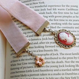 May include: A pink velvet ribbon bookmark with a gold chain and a cameo pendant. The cameo is a red and gold oval with a white profile of a woman's face. The text on the page reads "Scotsman and New York. ... the first time he had ... the continuity and ... to a less frenetic life."