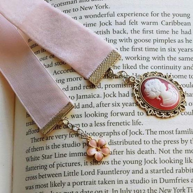 May include: A pink velvet ribbon bookmark with a gold chain and a cameo pendant. The cameo is a red and gold oval with a white profile of a woman's face. The text on the page reads "Scotsman and New York. ... the first time he had ... the continuity and ... to a less frenetic life."