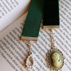 May include: A green velvet bookmark with a silver chain and a cameo pendant featuring a woman's profile. The cameo is set in a silver frame with a pearl-like embellishment.