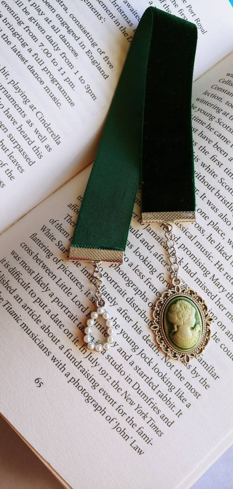 May include: A green velvet bookmark with a silver chain and a cameo pendant featuring a woman's profile. The cameo is set in a silver frame with a pearl-like embellishment.