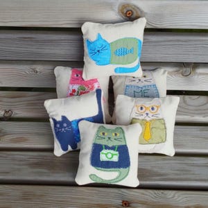 May include: Five small, handmade cat-shaped pillows with different designs. The pillows are made of white fabric with colorful embroidered details. One pillow features a blue cat with a green fish, another has a pink cat with flowers, another has a gray cat with a blue sweater, another has a white cat with glasses and a yellow tie, and the last one has a green cat with a blue shirt and a camera.