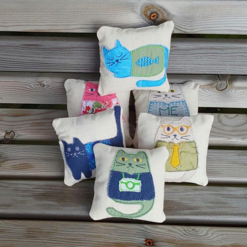 May include: Five small, handmade cat-shaped pillows with different designs. The pillows are made of white fabric with colorful embroidered details. One pillow features a blue cat with a green fish, another has a pink cat with flowers, another has a gray cat with a blue sweater, another has a white cat with glasses and a yellow tie, and the last one has a green cat with a blue shirt and a camera.