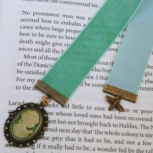 May include: A green velvet bookmark with a gold metal charm featuring a butterfly design. The bookmark is attached to a book with the text 'No prominent man was...' and 'Most of those who live...' visible.