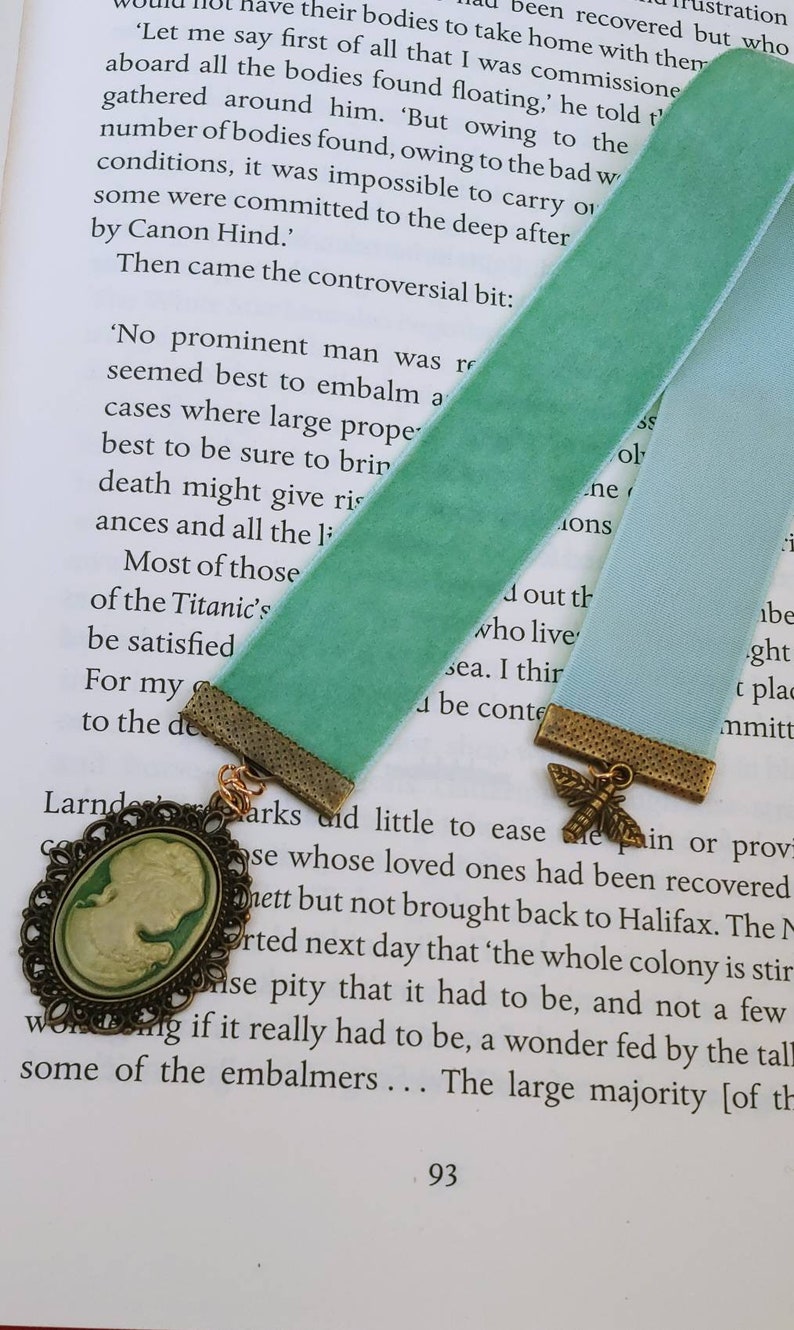 May include: A green velvet bookmark with a gold metal charm featuring a butterfly design. The bookmark is attached to a book with the text 'No prominent man was...' and 'Most of those who live...' visible.