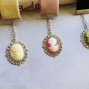 May include: Three vintage-style bookmarks with a cameo design. Each bookmark features a different colored cameo with a white profile of a woman. The bookmarks are attached to a ribbon and a chain.