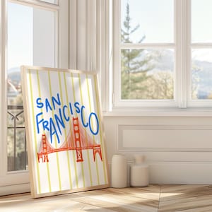 Puede incluir: Una impresión artística enmarcada con el puente Golden Gate en San Francisco. La obra de arte tiene un fondo blanco con rayas verticales amarillas. El puente está representado en naranja con texto azul "SAN FRANCISCO".