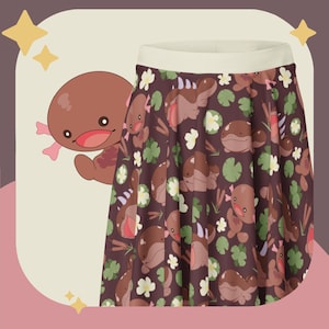 Puede incluir: Una falda acampanada con un patrón repetido de ajolotes de dibujos animados, nenúfares y flores sobre un fondo marrón oscuro. La falda tiene una cintura de color crema. Un ajolote de dibujos animados está en el lado izquierdo de la imagen. Es un artículo de moda divertido y caprichoso.