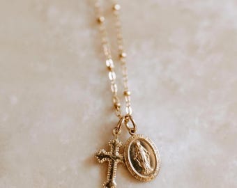 May include: A gold necklace featuring a delicate chain and two pendants. One pendant is a cross, and the other is an oval with a religious figure. The necklace is presented on a light-coloured surface.