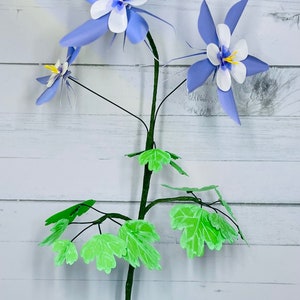May include: A paper craft bouquet of blue and white columbine flowers with green leaves. The flowers have a yellow center and are arranged on a black stem.