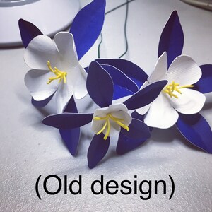 May include: Three white paper flowers with yellow centers and blue paper leaves. The flowers are arranged in a bouquet and the text "(Old design)" is visible below the flowers.