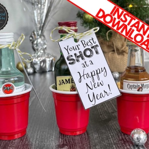 Puede incluir: Tres vasos de pl&aacute;stico rojos con botellas de licor en miniatura en su interior. Cada vaso tiene una etiqueta. La etiqueta del vaso del medio dice "Your Best Shot at a Happy New Year!"