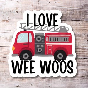 Op de afbeelding: Een sticker met een rode brandweerwagen met een ladder en de tekst "I LOVE WEE WOOS" in het zwart. De sticker heeft een witte rand en staat op een houten achtergrond.