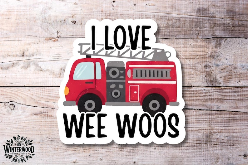Op de afbeelding: Een sticker met een rode brandweerwagen met een ladder en de tekst "I LOVE WEE WOOS" in het zwart. De sticker heeft een witte rand en staat op een houten achtergrond.