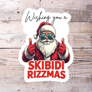 May include: A white sticker with a cartoon image of Santa Claus wearing sunglasses and giving a thumbs up. The text "Wishing you a Skibidi Rizzmas" is printed in red letters.