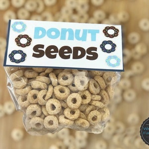 May include: A clear plastic bag filled with donut-shaped cereal. The bag has a blue and white label that says "Donut Seeds" with a donut graphic on each side.