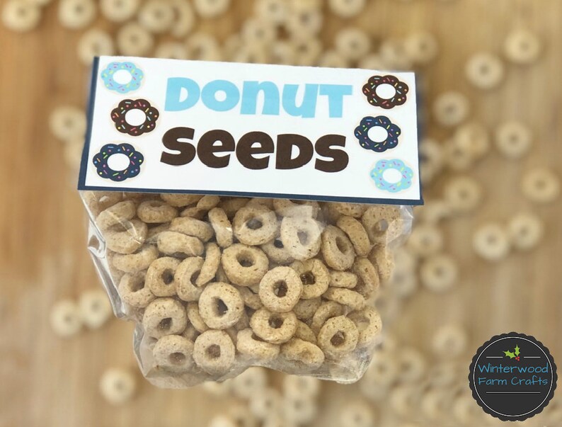 May include: A clear plastic bag filled with donut-shaped cereal. The bag has a blue and white label that says "Donut Seeds" with a donut graphic on each side.