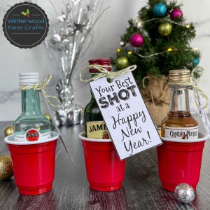 Puede incluir: Tres botellas de licor en miniatura en vasos de chupito de pl&aacute;stico rojo con una etiqueta de regalo con tem&aacute;tica navide&ntilde;a que dice "Your Best Shot at a Happy New Year!"