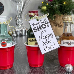 Puede incluir: Tres botellas de licor en miniatura en vasos de chupito de pl&aacute;stico rojo con una etiqueta de regalo con tem&aacute;tica navide&ntilde;a que dice "Your Best Shot at a Happy New Year!"