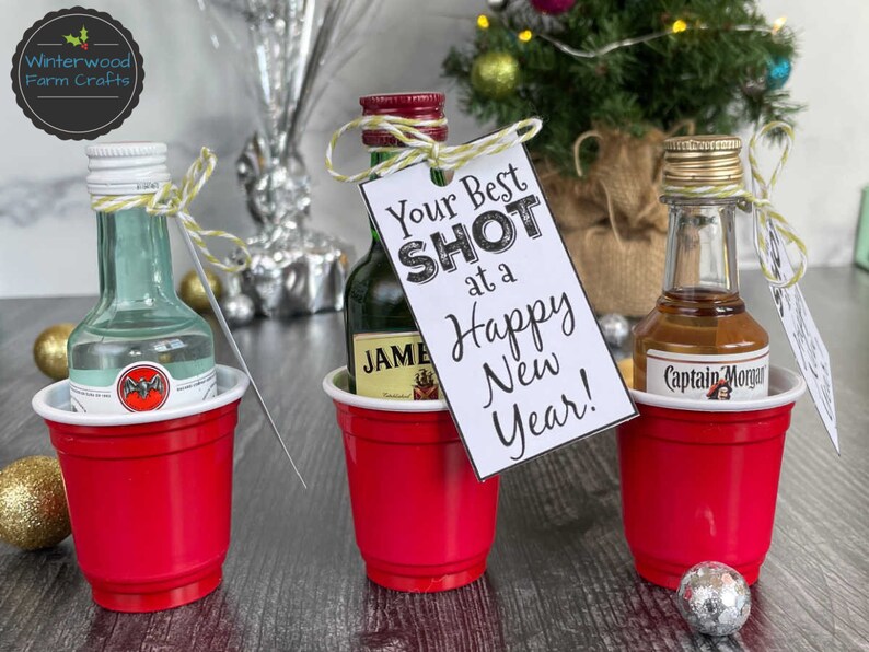 Puede incluir: Tres botellas de licor en miniatura en vasos de chupito de pl&aacute;stico rojo con una etiqueta de regalo con tem&aacute;tica navide&ntilde;a que dice "Your Best Shot at a Happy New Year!"