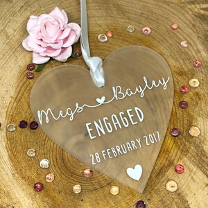 May include: A frosted heart-shaped acrylic ornament with the text "Megs & Bayley ENGAGED 28 FEBRUARY 2017" in white lettering. A small white heart is at the bottom of the ornament.