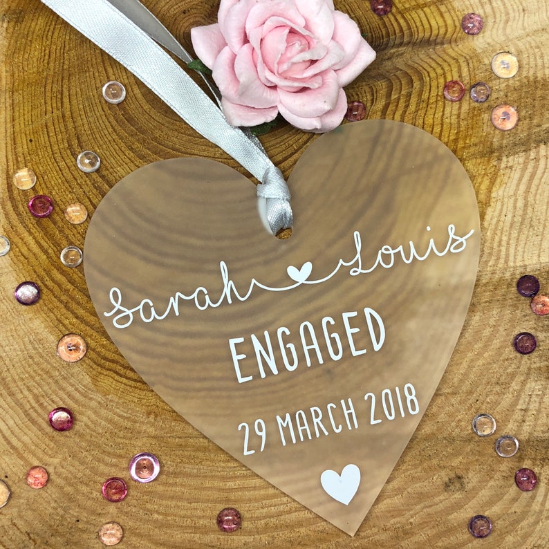 May include: A clear heart-shaped acrylic ornament with the text "Sarah & Louis Engaged 29 March 2018" in white lettering.