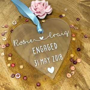 May include: A clear heart-shaped acrylic ornament with the text "Rosie & Craig ENGAGED 31 MAY 2018" in white lettering.