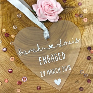 May include: A clear heart-shaped acrylic ornament with the text "Sarah & Louis Engaged 29 March 2018" in white lettering.