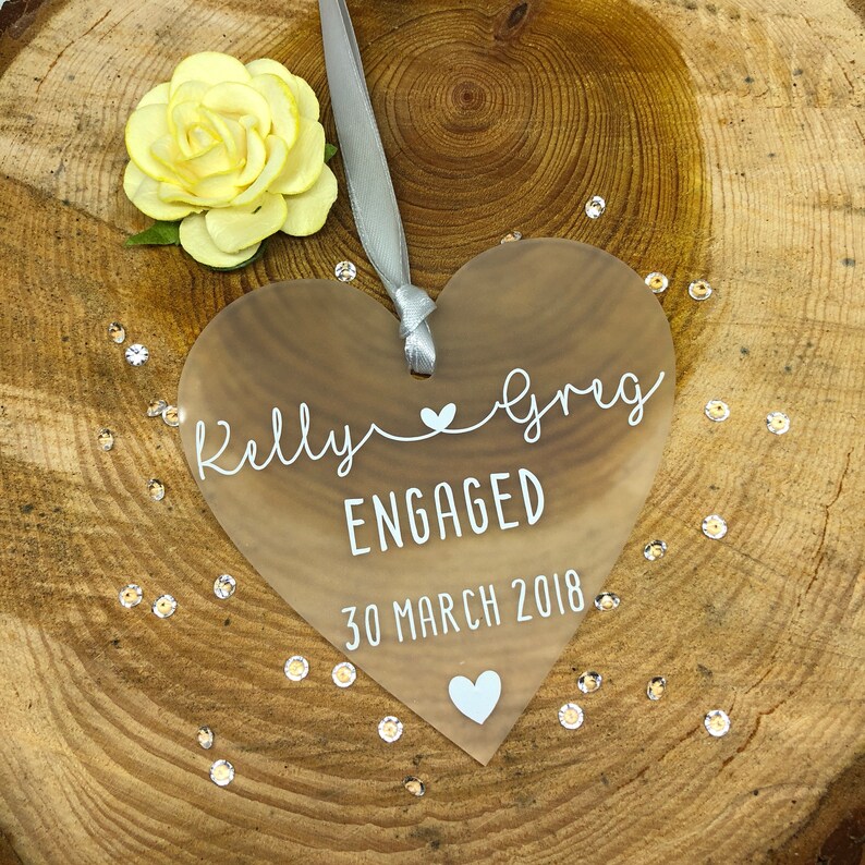May include: Clear heart-shaped acrylic ornament with the text "Kelly & Greg Engaged 30 March 2018" and a small heart symbol.