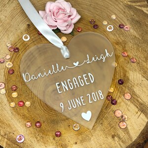 May include: A frosted clear heart-shaped acrylic ornament with the text "Danielle & Sleigh ENGAGED 9 JUNE 2018" in white lettering.