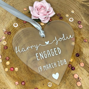May include: A frosted heart-shaped acrylic ornament with the text "Harry & Jodie ENGAGED 19 March 2018" in white lettering.
