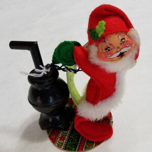May include: A vintage felt Santa Claus doll holding a green sack and standing next to a black wood stove. The doll is wearing a red hat with green holly, a red suit, and white fur trim. The doll is standing on a round base with a colorful patterned fabric.