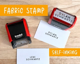 May include: A red self-inking fabric stamp with the text "DYLAN SCHWARTZ" on the top and "printy Clothing Marker" on the bottom. The stamp is on a white card with the text "DYLAN SCHWARTZ" printed on it. There is a second white card with the text "LEXI SCHWARTZ" printed on it. The text "SELF-INKING" is printed in orange on a white background.
