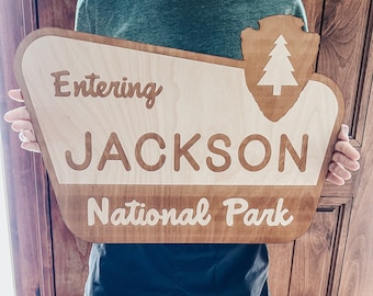 May include: A wooden sign with a brown stain and white lettering. The sign reads "Entering Jackson National Park" and features a stylized tree inside a shield shape.