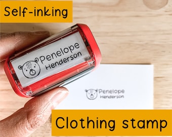 May include: Red self-inking clothing stamp with a white label that says "Penelope Henderson" and a small bear illustration. The stamp is being used to create an imprint on a white card.