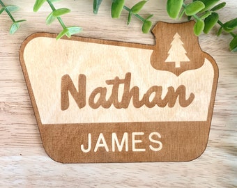 May include: Wooden name sign with the name "Nathan" in a brown script font and the last name "JAMES" in a block font. The sign has a light wood background and a dark wood border with a tree graphic.