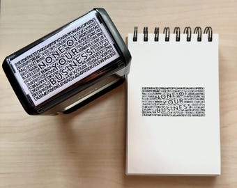May include: A black stamp with the words "NONE OF YOUR BUSINESS" printed on it, next to a white notepad with the same text stamped on it. The stamp is angled, showing the text in a rectangular shape.