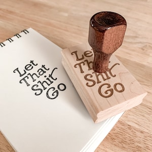 May include: Wooden stamp with the text "Let That Shit Go" in a cursive font. The stamp is made of light wood and has a dark brown wooden handle.