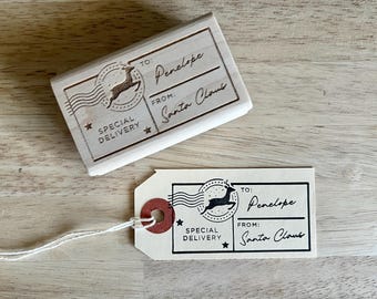 May include: Two wooden stamps and a tag with the text "TO: Penelope FROM: Santa Claus" and "SPECIAL DELIVERY". The stamps feature a reindeer design inside a circle with stars. The tag has a red circular reinforcement.