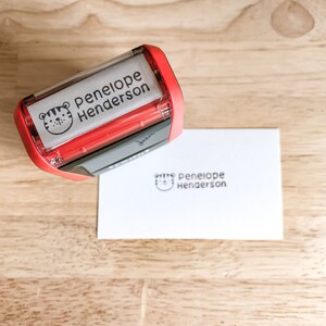 Pu&ograve; includere: Timbro autoinchiostrante rosso e nero con un'icona di viso di tigre e il testo "Penelope Henderson". Il timbro viene utilizzato per creare un'impressione di inchiostro nero dello stesso testo e icona su un cartoncino bianco.
