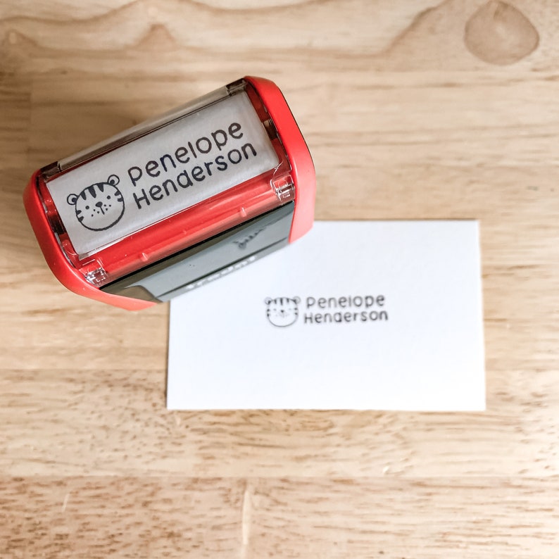 Pu&ograve; includere: Timbro autoinchiostrante rosso e nero con un'icona di viso di tigre e il testo "Penelope Henderson". Il timbro viene utilizzato per creare un'impressione di inchiostro nero dello stesso testo e icona su un cartoncino bianco.