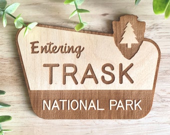 May include: Wooden sign with the text "Entering Trask National Park" in a vintage style font. The sign has a brown woodgrain finish and a stylized tree icon in the upper right corner.