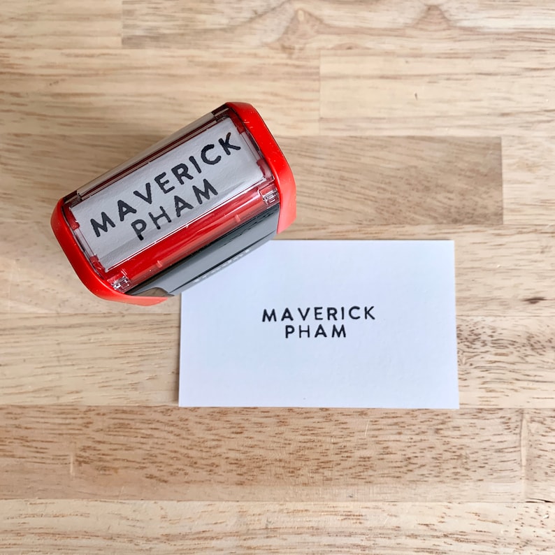 May include: A red self-inking stamp with the text "MAVERICK PHAM" in black lettering. The stamp is on a wooden surface and next to a white card with the same text printed on it.