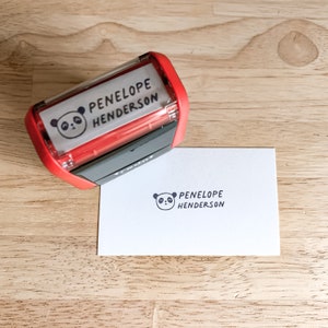 Pu&ograve; includere: Timbro autoinchiostrante rosso e nero con un viso di panda e il testo "PENELOPE HENDERSON". Il timbro viene utilizzato per creare un'impressione di inchiostro nero su un cartoncino bianco.
