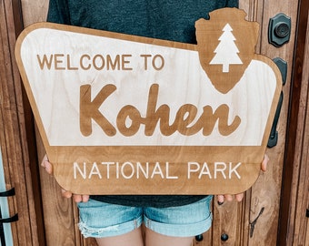 May include: A wooden sign with the text "Welcome to Kohen National Park" in white lettering. The sign is brown with a white silhouette of a pine tree in the upper right corner.