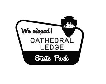 Könnte beinhalten: Ein schwarz-weißes grafisches Schild mit dem Text We eloped! CATHEDRAL LEDGE State Park. Das Design enthält ein stilisiertes Pfeilspitzen-Symbol mit einer Bergsilhouette, eingefasst in einen abgerundeten trapezförmigen Rahmen mit schwarzer Basis.