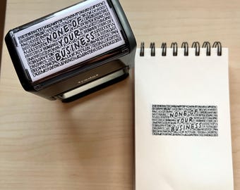 May include: A black self-inking stamp with the text "NONE OF YOUR BUSINESS" in a stylized font. A white notepad with the same text stamped on it is next to the stamp.