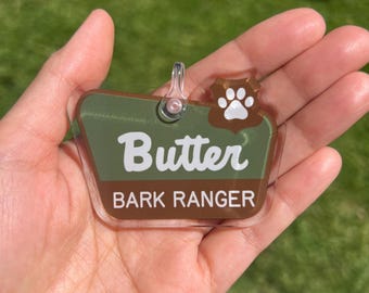May include: A clear acrylic pet tag shaped like a badge with a green and brown background. The tag features the white text Butter and BARK RANGER, along with a white paw print icon. It includes a small clear loop at the top for attachment.