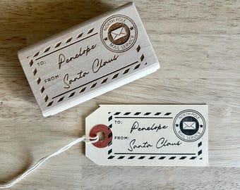 May include: A wooden stamp and a tag with the text "To: Penelope From: Santa Claus". The stamp has a North Pole Mail Service logo. The tag has a red reinforced hole and a string.