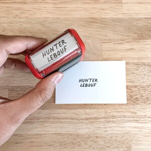 May include: A red self-inking stamp with the text "HUNTER LEBOUF" in black ink. The stamp is being used to stamp the same text onto a white card.